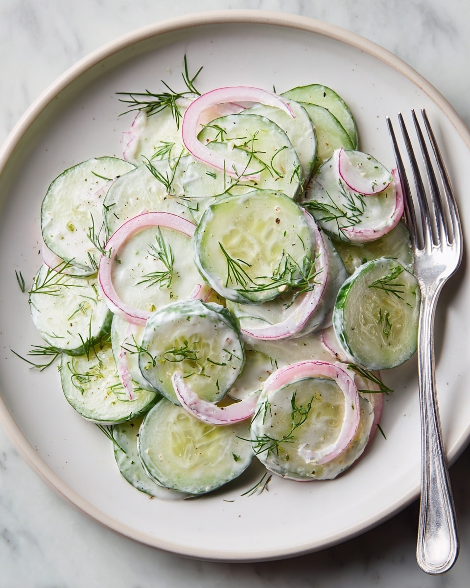 A white bowl holds a fresh cucumber salad made of around three layers of thinly sliced cucumber rounds with green edges and pale green centers, mixed evenly with thin slices of pale purple onion. The vegetables are coated in a creamy white dressing with a smooth texture, lightly dripped over each slice. Small green dill sprigs are scattered throughout the salad, adding detail and a touch of color contrast. A shiny silver spoon rests inside the bowl, partially covered with the salad. The bowl is placed on a white marbled surface. Photo taken with an iphone --ar 4:5 --v 7