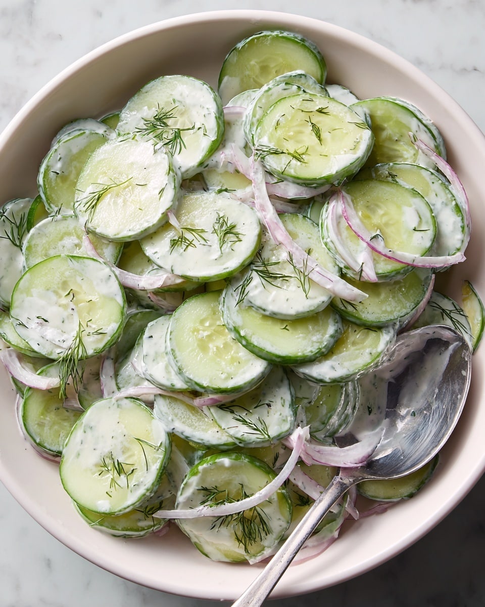 A white plate holds a fresh cucumber salad with about two layers of thinly sliced cucumbers coated lightly in a creamy white dressing. Thin slices of red onion are scattered among the cucumbers, adding a touch of pale pink color. Small green dill sprigs are sprinkled evenly across the salad, giving it a fresh and herbaceous look. A silver fork rests to the right side of the plate, all set against a white marbled surface. Photo taken with an iphone --ar 4:5 --v 7
