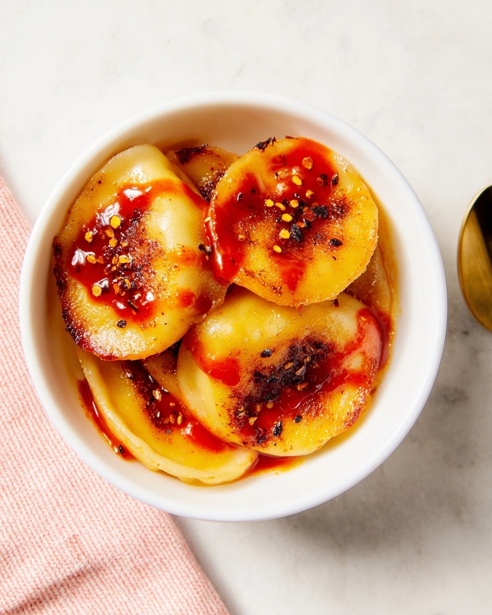 In a white bowl, there are three soft, golden yellow dumplings arranged close together. Each dumpling is topped with a bright red sauce drizzled unevenly across the surface, adding a glossy texture and a pop of color. Small black and red seasoning flakes are sprinkled over the dumplings, giving a speckled effect. The edges of the dumplings appear slightly crispy and browned in places, contrasting with the smooth, soft centers. The bowl sits on a white marbled surface. photo taken with an iphone --ar 4:5 --v 7