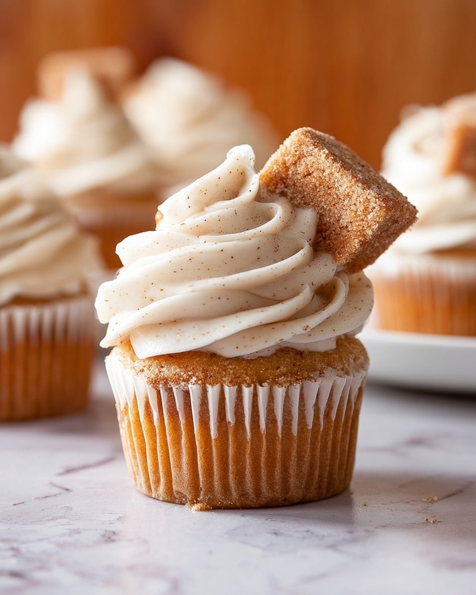 A single cupcake with a golden brown base wrapped in a white paper liner sits on a white marbled surface. On top, there is a thick swirl of smooth, creamy off-white frosting with tiny specks, giving it a soft texture. A small rectangular piece of cinnamon sugar-coated cake is gently placed at an angle, partially embedded in the frosting at the back. In the blurred background, more similarly frosted cupcakes are arranged on a white plate. photo taken with an iphone --ar 4:5 --v 7