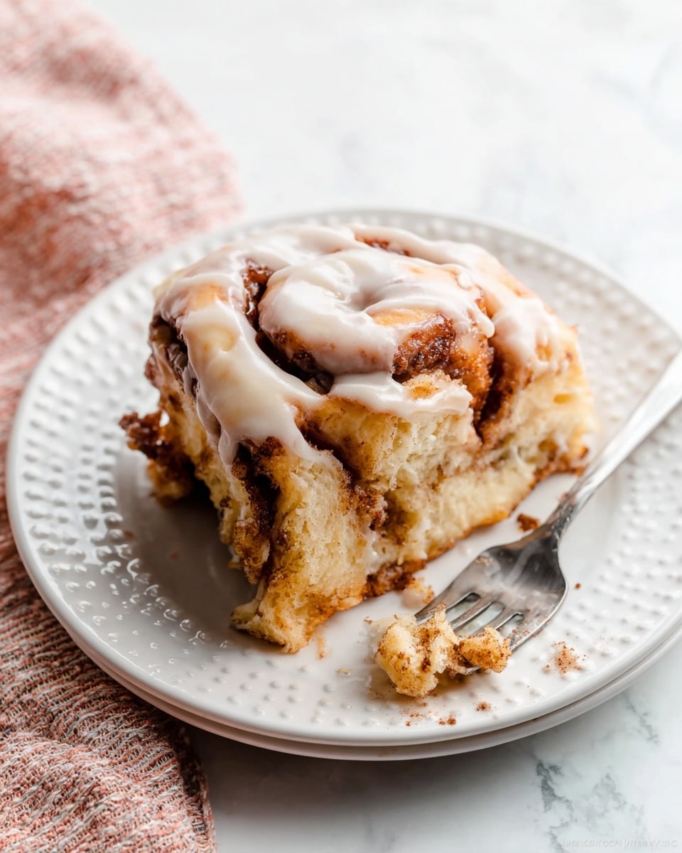 A slice of cinnamon roll sits on a white plate with small raised dot details around the edge. The roll has about four thick, soft layers, each showing a light golden brown dough with darker cinnamon swirls inside. The top of the roll is covered with a smooth and creamy white glaze that drips slightly down the sides. Crumbs and bits of cinnamon fill the space between layers. A silver fork with some cinnamon crumbs is placed near the bottom right on the plate. The plate rests on a white marbled surface with a soft pink and white woven cloth partially visible in the bottom left corner. Photo taken with an iphone --ar 4:5 --v 7