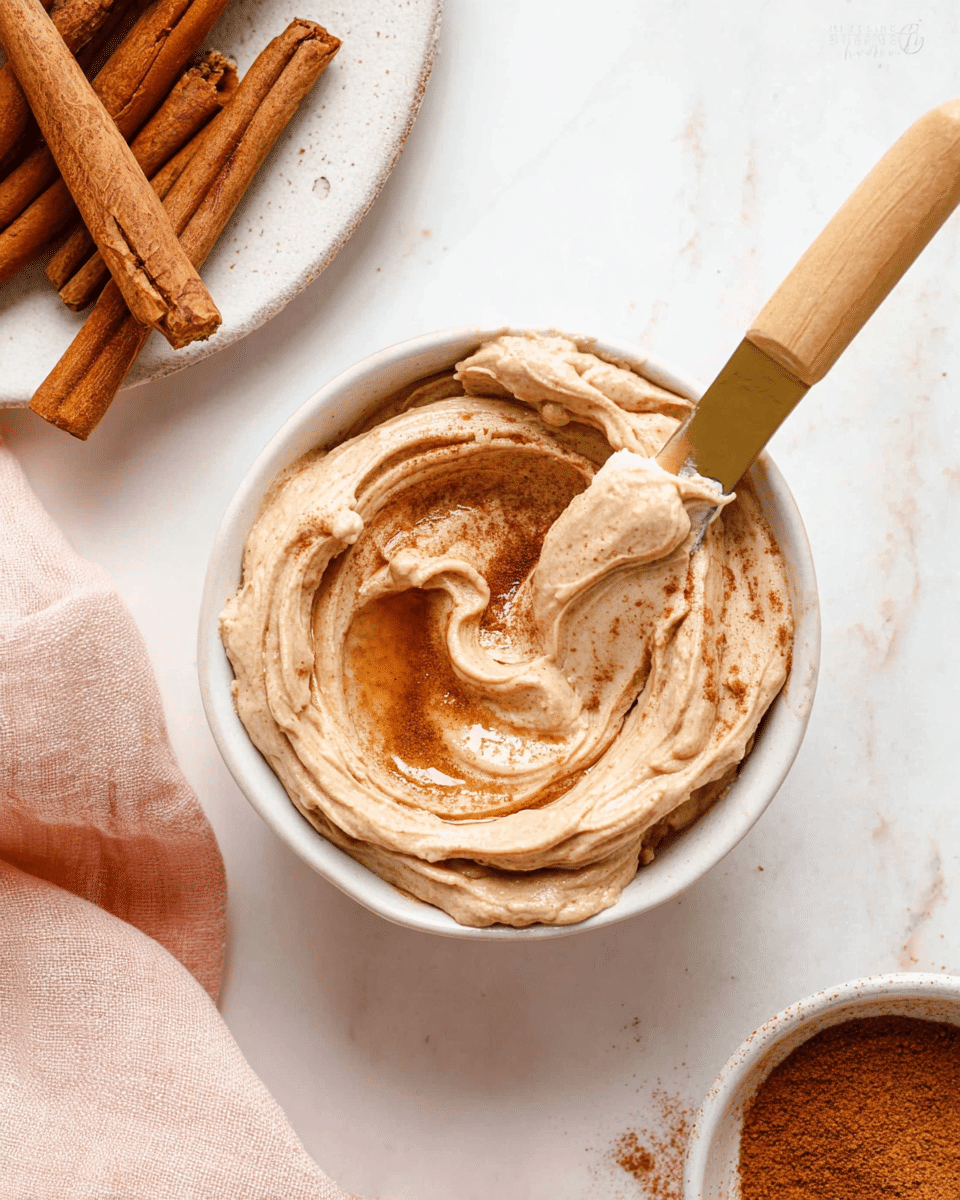 A small white bowl filled with a creamy light brown mixture swirled smoothly with a shiny honey-like layer in the middle sprinkled with fine darker brown cinnamon powder. A butter knife with a light wooden handle and a gold blade rests inside the bowl with some of the creamy spread on the blade. Near the bowl, there are several whole cinnamon sticks on a white plate with a rough edge, and part of a white bowl with more cinnamon powder visible. The setup is on a white marbled surface with a soft pink cloth partially visible in the corner. photo taken with an iphone --ar 4:5 --v 7