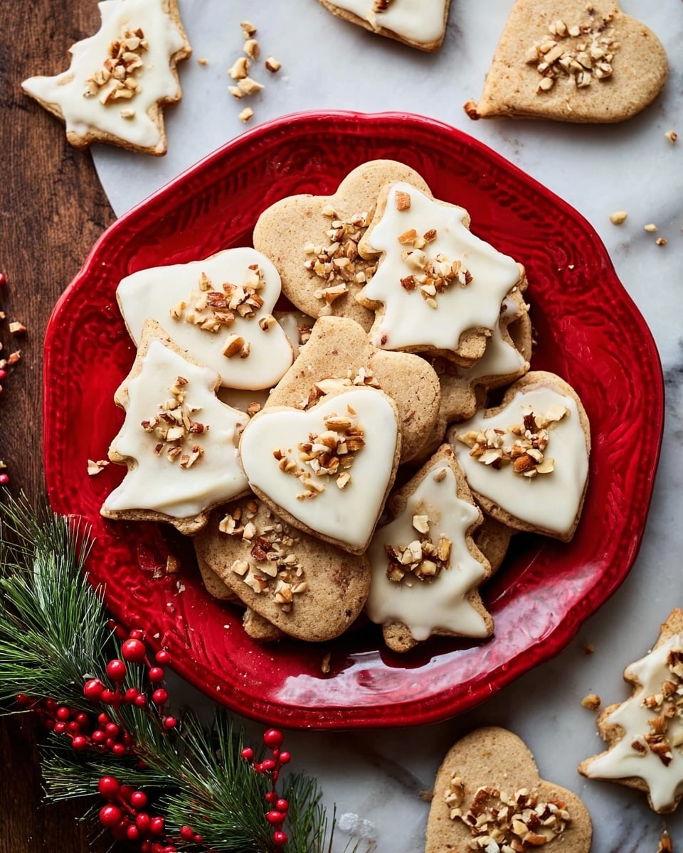 A red plate holds a pile of shaped cookies including hearts and Christmas trees, all light brown with bits of nuts inside. On top of the cookies is a smooth white icing layer, each cookie decorated with chopped nuts sprinkled over the icing. Around the plate, more shaped cookies are scattered on a white marbled surface, and there is a small sprig of pine with red berries near the bottom left. Photo taken with an iphone --ar 4:5 --v 7