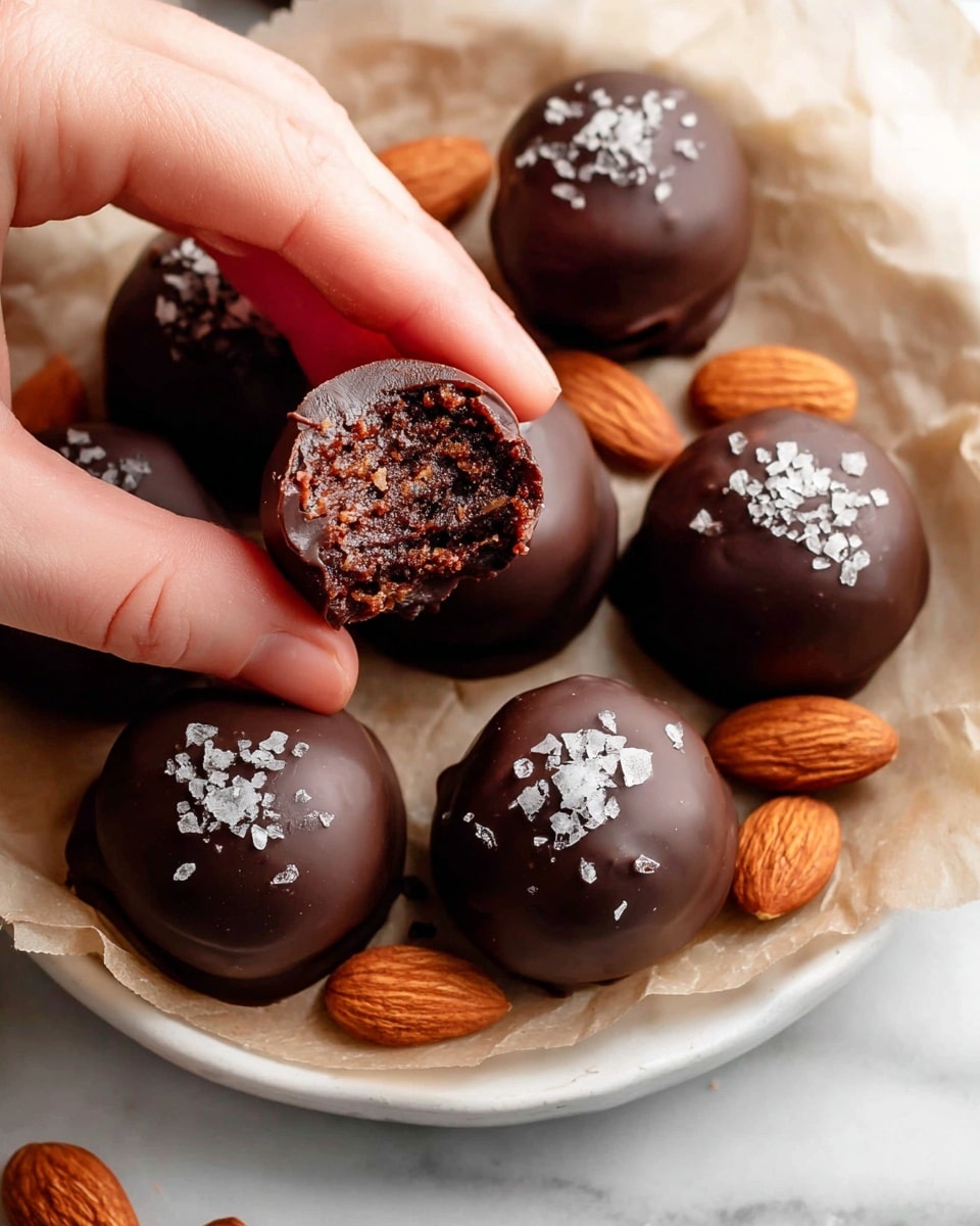 A close-up view of several small, round chocolate truffles with a smooth, dark brown shiny outer layer, topped with flakes of coarse sea salt. Inside each truffle, there is a dense, crumbly, dark filling visible in one truffle held by a woman's hand in the foreground, showing a textured mixture with tiny nut pieces. Whole almonds are scattered among the truffles on a crinkled parchment paper, all placed on a white plate with a slight scalloped edge. The background features a white marbled texture. Photo taken with an iphone --ar 4:5 --v 7