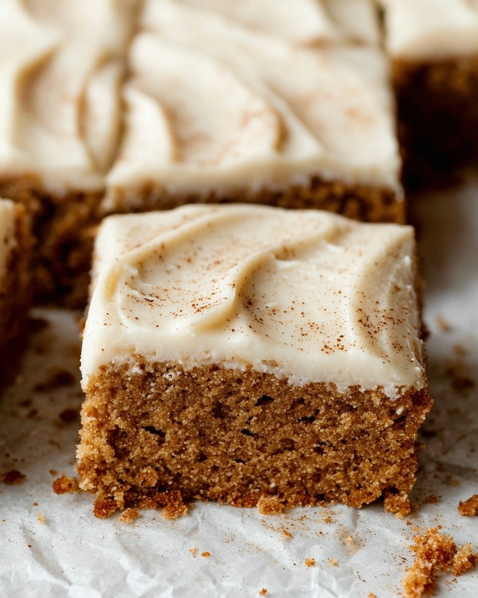 This close-up image shows a square piece of moist, brown cake with a crumbly texture, topped with a thick, smooth layer of creamy white frosting that has a few swirls and is lightly sprinkled with brown specks, resembling cinnamon or nutmeg. The cake is part of a larger batch cut into squares, resting on a white marbled textured surface with some crumbs scattered around. The frosting layer covers the entire top evenly and appears soft and rich. Photo taken with an iphone --ar 4:5 --v 7
