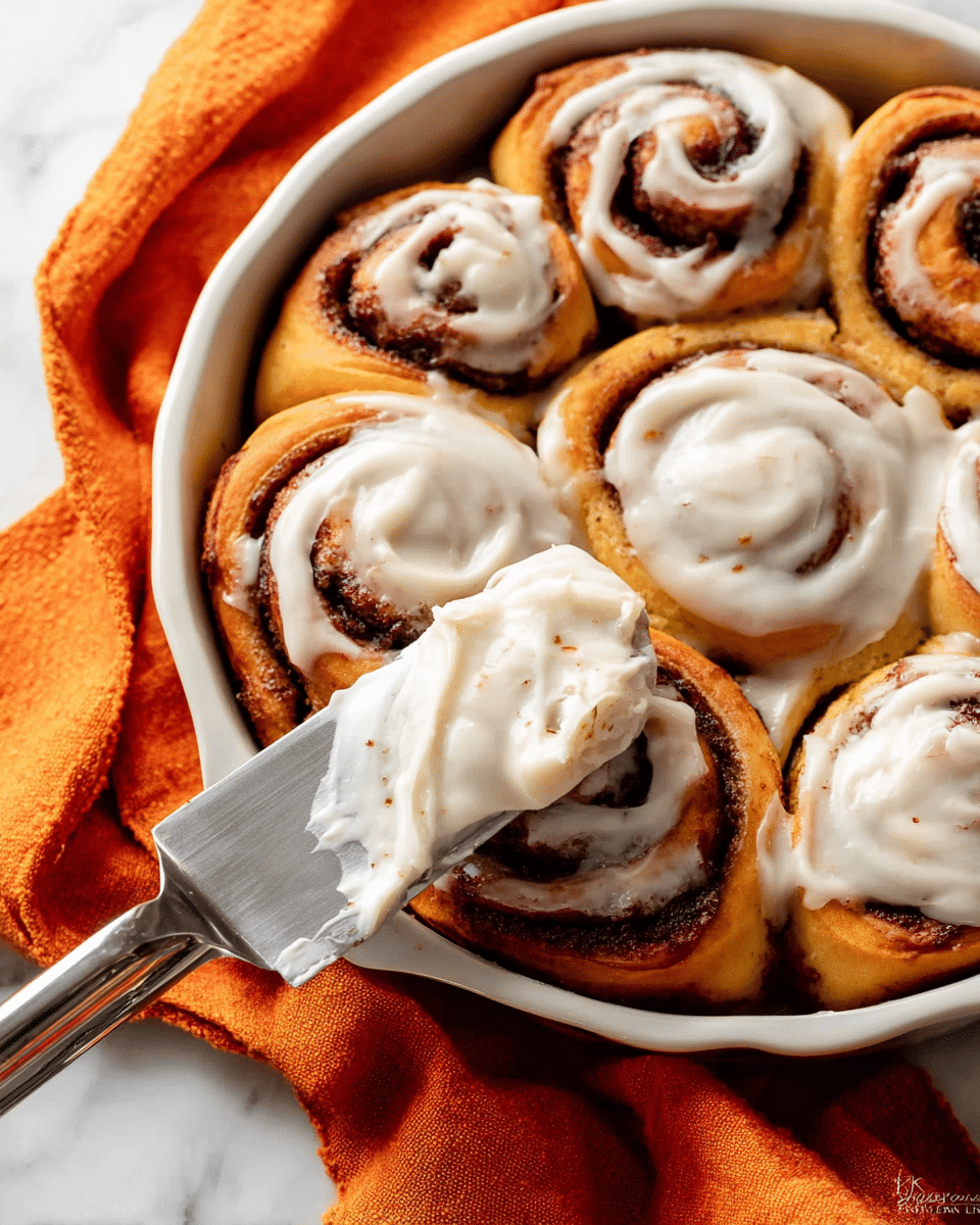 A close-up of a white ceramic dish filled with soft, golden brown cinnamon rolls arranged tightly in a single layer, each roll showing spirals of dark cinnamon filling. Some rolls are topped with thick, creamy white frosting that has visible tiny specks, spread unevenly on top. A metal spatula covered with frosting rests on one roll, partially spreading the icing. The dish sits on a bright orange cloth on a white marbled surface. Photo taken with an iphone --ar 4:5 --v 7