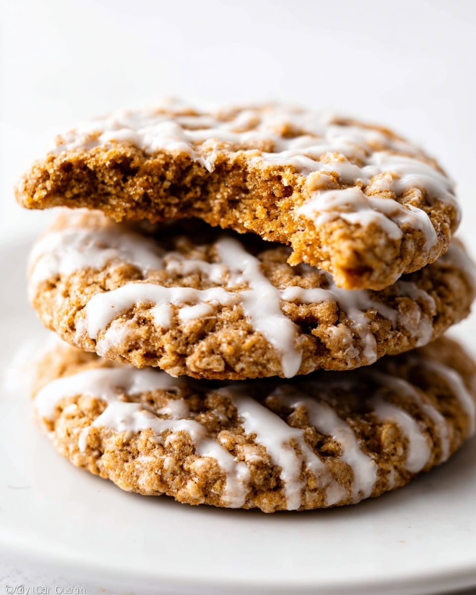 A close-up view of three round oatmeal cookies stacked on top of each other on a white plate, placed on a white marbled surface. Each cookie has a rough, grainy texture with a golden brown color. The top cookie is broken in half, showing a soft and crumbly inside that matches the golden brown outside. White icing is drizzled unevenly across each cookie in a pattern that covers parts of the cookie surface, adding contrast and a smooth, glossy texture against the roughness of the oatmeal base. The cookies fill most of the frame with the focus on their texture and icing details. Photo taken with an iphone --ar 4:5 --v 7