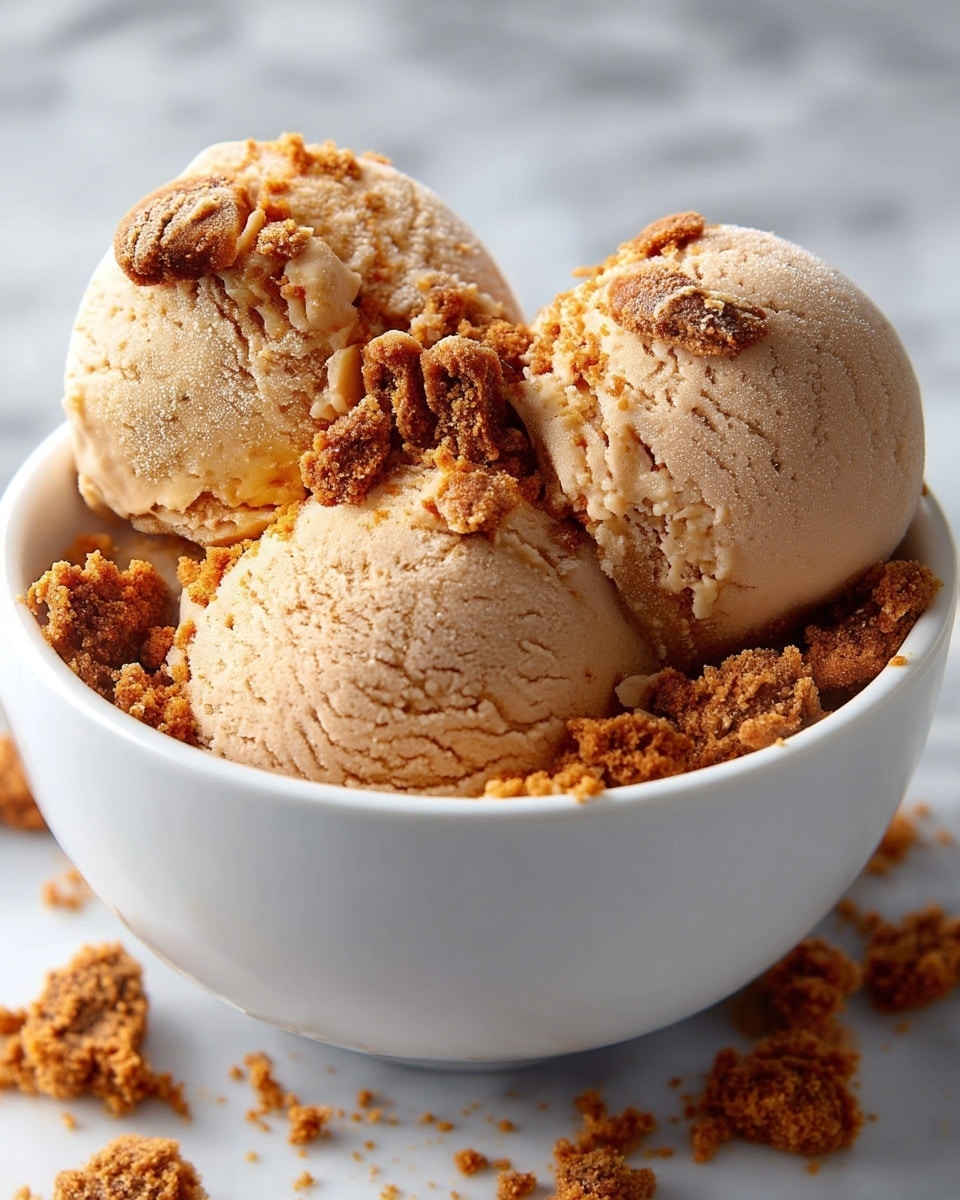 A close-up view of three scoops of smooth, light brown ice cream in a white bowl, with a crumbly, darker brown cookie or biscuit topping scattered unevenly on and around the ice cream scoops, adding a crunchy texture contrast. The background shows a soft, blurred white marbled surface. photo taken with an iphone --ar 4:5 --v 7