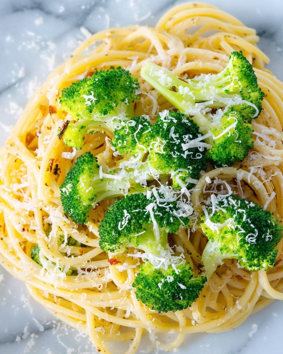This close-up image shows a dish with three main layers. The bottom layer is light yellow spaghetti noodles, slightly glossy and mixed with small bits of seasoning. On top of the noodles, there are bright green broccoli florets with a fresh texture, some cut to show a pale green inside. The top layer is sprinkled with fine white grated cheese, scattered unevenly across the broccoli and noodles, creating a light snowy effect. The whole dish rests on a white marbled surface. photo taken with an iphone --ar 4:5 --v 7
