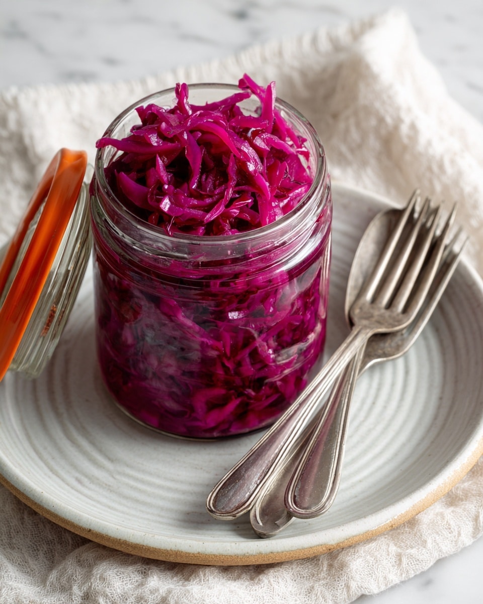A glass jar filled with bright purple-red shredded pickled cabbage sits open on a white plate with a textured rim. Two vintage metal forks rest on the plate next to the jar. The jar has an orange rubber seal around its lid, which is open and leaning to the side. The whole scene is set on a white marbled surface with a soft off-white cloth nearby, adding a cozy touch. The pickled cabbage inside the jar looks wet and shiny, with thin strands packed together closely. photo taken with an iphone --ar 4:5 --v 7