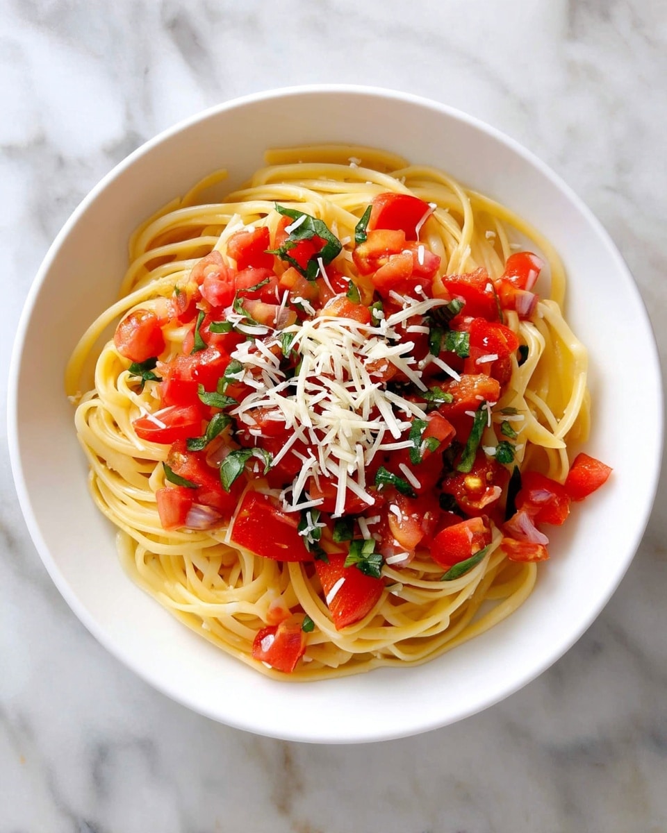 A white bowl filled with a single layer of spaghetti pasta as the base, light yellow in color with a smooth, slightly oily texture. On top, there is a generous layer of small, bright red diced tomatoes mixed with finely chopped green basil leaves, adding a fresh, vibrant look. Scattered over the tomatoes and pasta are thin shreds of white cheese, offering a soft, slightly crumbly texture. The bowl sits on a white marbled surface. photo taken with an iphone --ar 4:5 --v 7