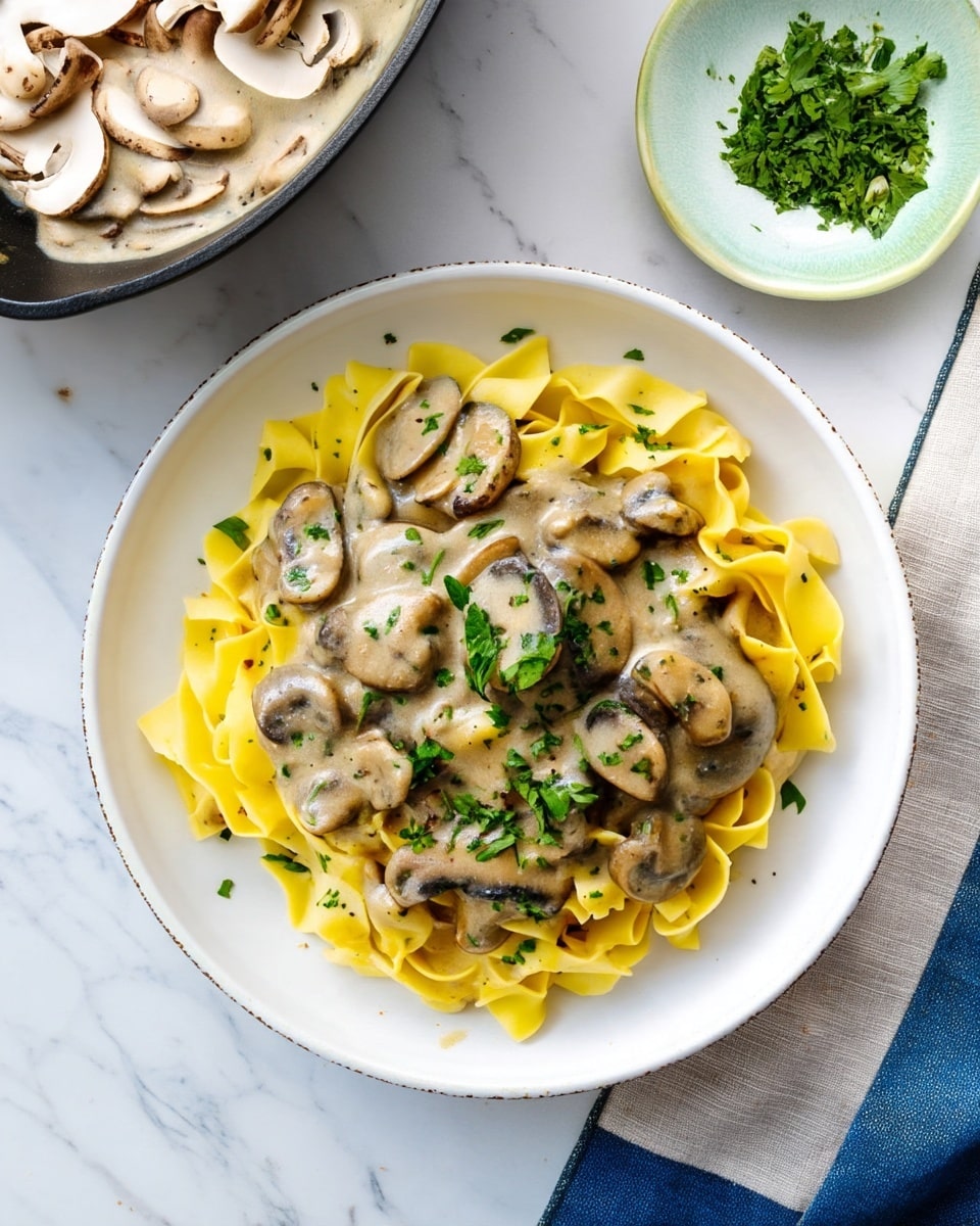 A white round plate holds a serving of wide yellow pasta ribbons, loosely folded. On top, there is a thick creamy mushroom sauce with light brown and gray sliced mushrooms scattered across the pasta. The sauce has a smooth texture with some herbs mixed in. Fresh chopped green herbs are sprinkled over the dish for garnish. The plate is set on a white marbled surface with a small white bowl filled with chopped green herbs to the top right and a gray pan containing mushroom sauce to the top left. At the bottom left, part of a white bowl with thinly sliced mushrooms is visible, and a beige cloth with a blue edge lies at the bottom right. Photo taken with an iphone --ar 4:5 --v 7