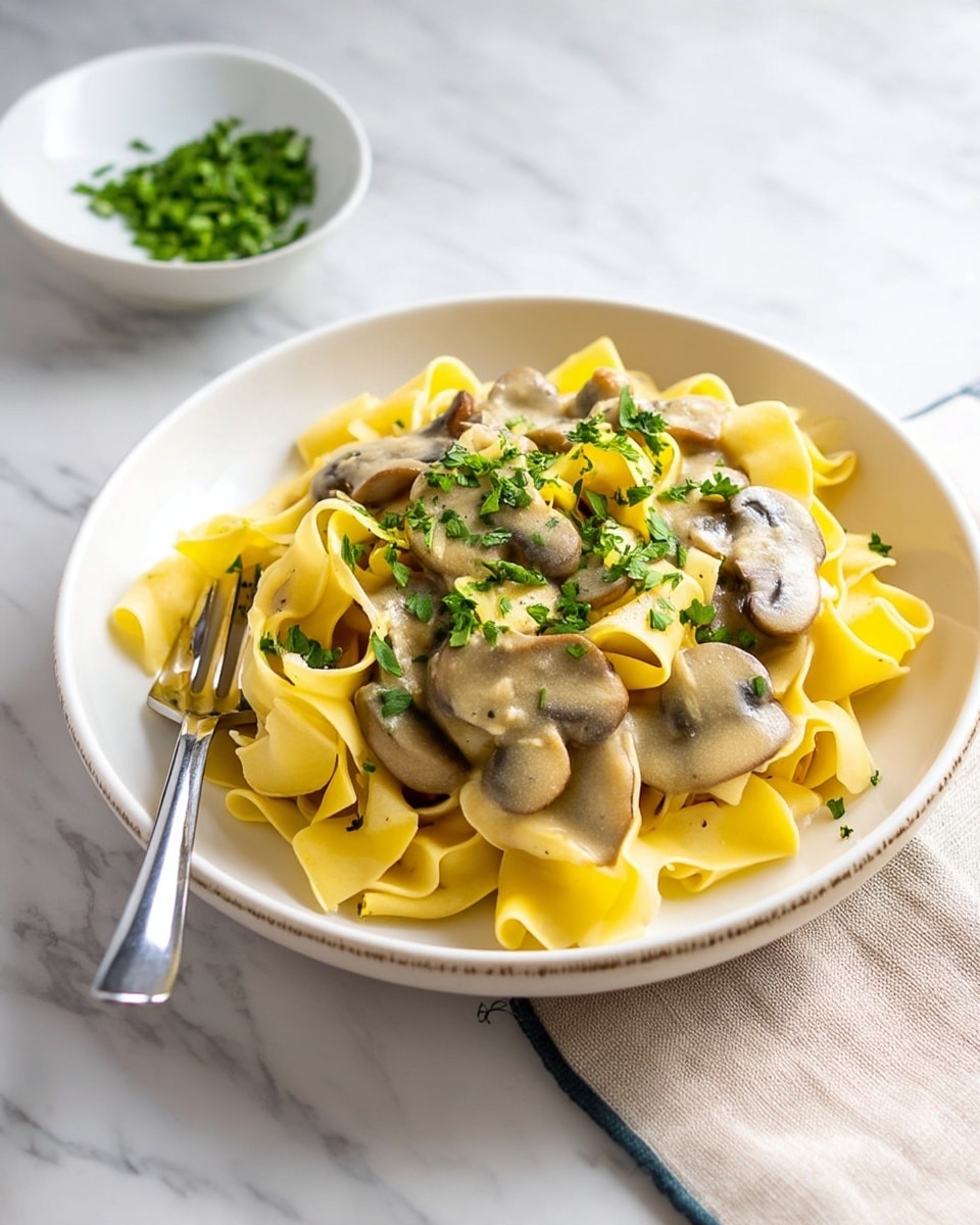 A white plate holds a serving of wide, yellow pasta noodles layered loosely in a pile. On top, sliced mushrooms covered in a creamy, glossy light beige sauce spread unevenly across the pasta. Bright green chopped herbs are sprinkled over the dish, adding a touch of color contrast. A silver fork rests on the edge of the plate. In the background, there is a small white bowl with finely chopped green herbs. The whole scene is set on a white marbled surface with a beige cloth napkin at the corner. photo taken with an iphone --ar 4:5 --v 7
